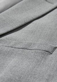 Grauer Stoff in Nahaufnahme mit glatter Textur, subtilem Muster und einer Paspeltasche oben links.