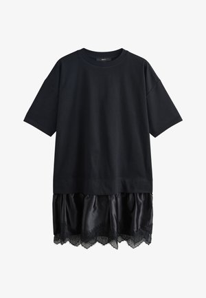 Vrouw met haar in een knot, gekleed in een zwart oversized overhemd en een rok met kanten rand, draagt een zwart-wit patroon schoudertas, van achteren gezien.
