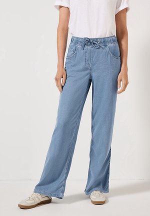 Personne portant un pantalon en jean bleu clair à jambes larges avec une taille élastique et un cordon, associé à des baskets blanches et un t-shirt blanc.