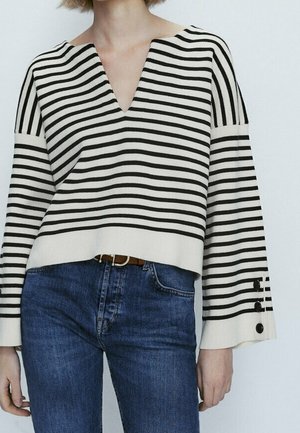Frau trägt einen schwarz-weiß horizontal gestreiften V-Ausschnitt-Pullover mit weiten Ärmeln und blaue Denim-Jeans.