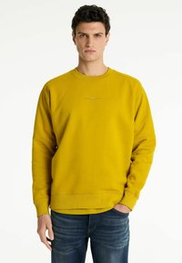 Sudadera amarilla de manga larga, con cuello redondo acanalado y un sutil logo en el pecho. Presenta un corte relajado y una textura de tela. Se combina con jeans azules.