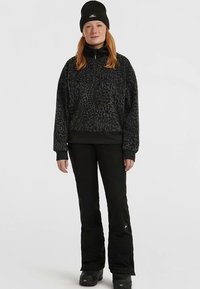 Zwarte fleece pullover met luipaardprint, kwartritssluiting bij de hals en zwarte accenten, gecombineerd met zwarte broek en een gebreide beanie.