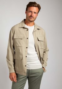 Beige lichte jas met knoopsluiting, vier voorzakken, een kraag en een rechte snit, gedragen over een witte shirt en groene broek.