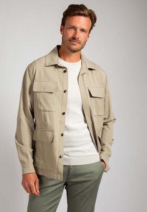 Beige lightweight jacket with button closure, four front pockets, a collar, and a straight cut, worn over a white shirt and green pants.
