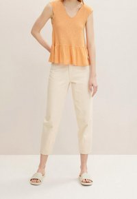 Woman wearing sleeveless orange ruffled top, off-white high-waist cropped pants, and beige quilted slide sandals, standing against plain background.
