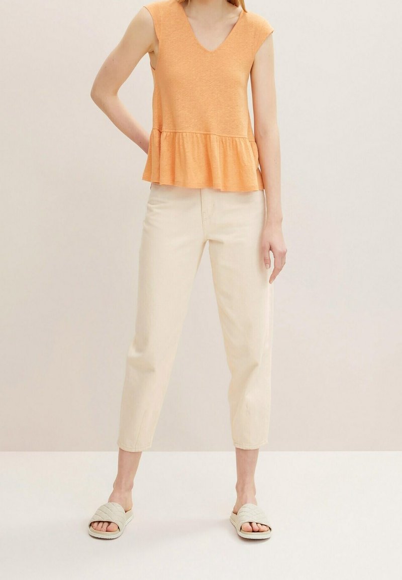 Woman wearing sleeveless orange ruffled top, off-white high-waist cropped pants, and beige quilted slide sandals, standing against plain background.