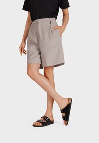 Shorts beige a vita alta con pieghe e tasche, abbinati a sandali neri senza chiusura con due fibbie e una soletta in sughero.