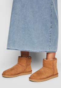 Stivaletti alla caviglia in suede beige con fodera morbida, punta curvata e suola testurizzata, abbinati a una gonna di jeans azzurro chiaro. Visibili dettagli delle cuciture.