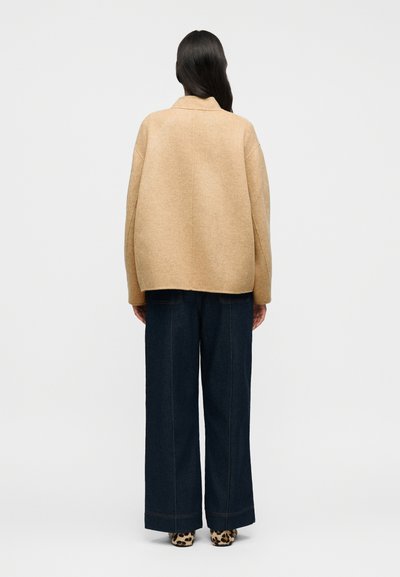 Μπεζ μάλλινο υπερμεγέθες σακάκι, άνετη εφαρμογή, με λεπτομέρεια ραφής στην πλάτη, συνδυασμένο με παντελόνια φαρδιάς γραμμής από σκούρο τζιν και παπούτσια με ζωικό μοτίβο.