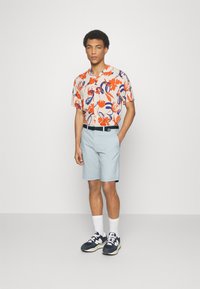 Chemise à manches courtes avec imprimé floral orange et bleu sur fond clair, associée à un short bleu clair, une ceinture noire et des baskets navy.