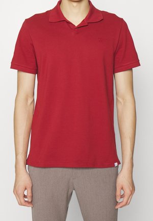 Mann trägt ein schlichtes rotes kurzärmeliges Poloshirt und hellbraune Hose, steht vor einem neutralen Hintergrund.