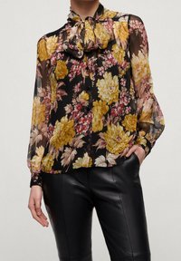 Blusa nera trasparente con motivi floreali gialli e rosa, scollo rotondo con nodo, chiusura a bottoni davanti e maniche lunghe con polsini aderenti.
