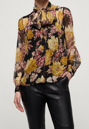 Transparante zwarte blouse met gele en roze bloemenpatronen, ronde halslijn met strik, knoopsluiting aan de voorkant en lange mouwen met nauwsluitende manchetten.