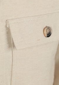 Beige stoffen overhemddetail met een voorzakje en een bijpassende knop. De textuur is glad met een subtiel geweven patroon.