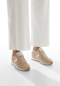 Baskets en daim beige avec un accent de talon blanc, des lacets en tissu et un logo réfléchissant. Assorties à un pantalon en velours côtelé blanc.