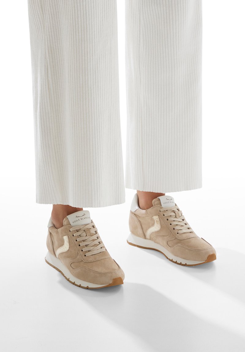Zapatillas de ante beige con un acento en el talón blanco, cordones de tela y un logo reflectante. Combinadas con pantalones de pana blanca.