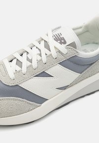 Chaussure de sport grise et blanche avec un mélange de daim, de mailles et de matériaux texturés. Les détails comprennent un logo et des lacets plats blancs.