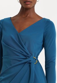 Blå långärmad klänning med V-ringning och snodd tygdetalj vid midjan, som hålls på plats av ett guldpläterat cirkelring. Slät textur.