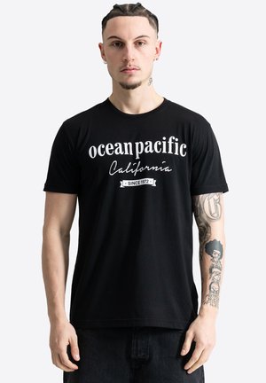 Hombre con el pelo trenzado que lleva una camiseta negra con la inscripción "ocean pacific California since 1972", mostrando el antebrazo tatuado sobre un fondo claro y liso.