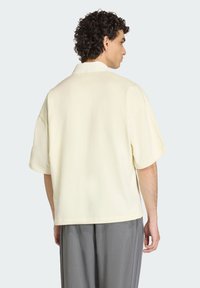 Koszula z krótkim rękawem w jasnym żółtym kolorze o luźnym kroju, lekko fakturowanej tkaninie, z pojedynczą kieszenią i otwartym kołnierzem. W zestawieniu z szarymi spodniami.