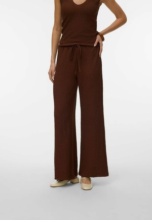 Femme portant un haut marron texturé sans manches et un pantalon à jambes larges avec des chaussures couleur crème, debout une main sur la hanche.