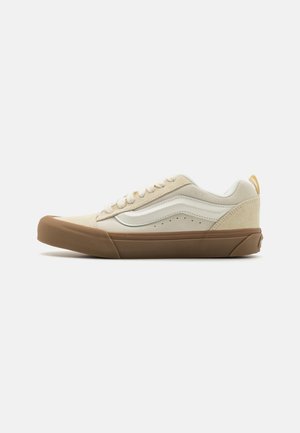 Baskets en toile beige avec des accents en suède, une bande en cuir blanc et une semelle en caoutchouc marron. Elles disposent de lacets ronds et de détails perforés sur le côté.