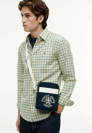 Joven con camisa a cuadros amarilla y azul y un bolso bandolera de Tommy Jeans en azul marino y blanco, posando frente a un fondo liso.