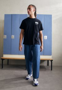 Tricou negru cu mânecă scurtă, cu text și grafică albă, asortat cu blugi de denim ușor albaștri și pantofi sport multicolori.