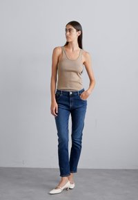 Μπέζ ραμμένο αμάνικο μπλουζάκι, μπλε τζιν παντελόνι με στενή εφαρμογή και λευκά τακούνια. Το φόντο είναι σε ουδέτερο γκρι.