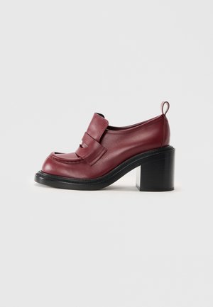 Mocasines de mujer en cuero rojo oscuro con tacón grueso negro y una cinta en la parte trasera, exhibidos sobre un fondo blanco liso.