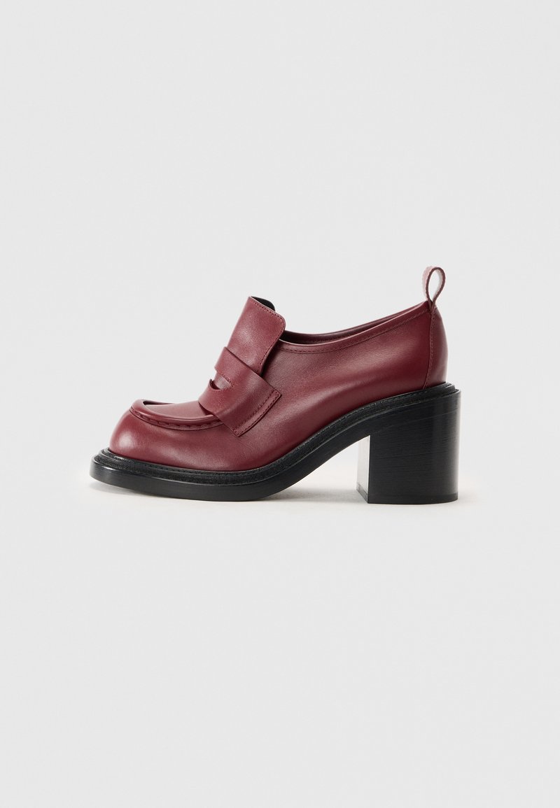 Mocasines de mujer en cuero rojo oscuro con tacón grueso negro y una cinta en la parte trasera, exhibidos sobre un fondo blanco liso.