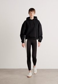 Felpa nera oversize, leggings neri con pannelli laterali bianchi e sneakers bianche con suola beige. Tessuto liscio, design minimalista.