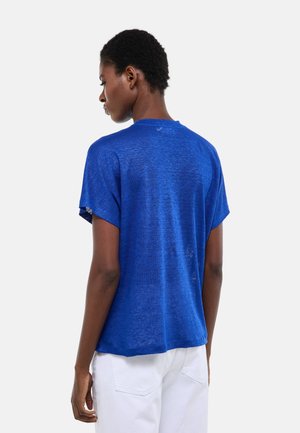 Femme aux cheveux courts portant une chemise bleue ample à manches courtes et un pantalon blanc, debout de dos sur un fond uni.