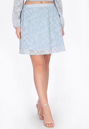 Kvinde iført en knælængde lysblå mønstret nederdel og sølvfarvede højhælede sandaler mod en hvid baggrund.