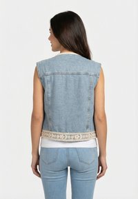 Mujer de espaldas que lleva un chaleco de mezclilla azul claro sin mangas con ribete de encaje floral beige sobre una camiseta blanca y vaqueros azules.