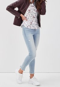 Veste en cuir marron, chemise blanche à fleurs à boutons, jeans skinny déchirés bleu clair et baskets blanches avec semelles texturées. Tenue décontractée.