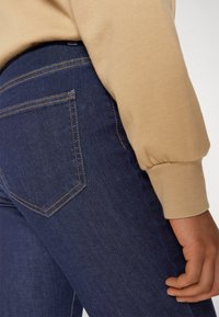 Tamsiai mėlyni džinsai su dviem galiniais kišenėmis, kontrastingos oranžinės siūlės, derinami su smėlio spalvos raižyto megztinio rankove.