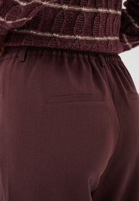 Gros plan d'une personne portant un pantalon taille élastique violet foncé avec des poches passepoilées à l'arrière et un pull en maille violet avec des rayures blanches.