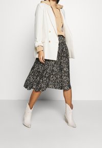 Witte blazer over een beige hoodie, gecombineerd met een zwarte bloemen midi-jurk en witte enkellaarsjes. Heeft een textuurstof en gelaagd ontwerp.