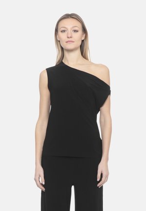 Donna che indossa un top nero monospalla e pantaloni neri, in piedi contro uno sfondo grigio chiaro uniforme, rivolta verso la fotocamera.