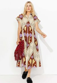Robe maxi crème avec des motifs géométriques en marron, violet et rouge. Comprend des manches courtes, un devant boutonné et un design fluide. Sac à franges rouges.