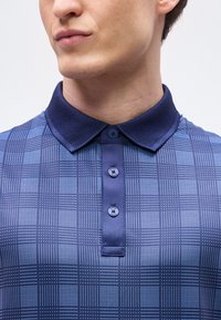 Κοντινό πλάνο ενός ατόμου που φοράει μπλε πόλο με σχέδιο, με υφασμένο σκούρο μπλε γιακά και τρία κουμπιά στο μπροστινό μέρος.