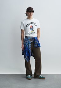 Hvit T-skjorte med grafisk trykk, blå og brun rutete skjorte knyttet rundt livet, brune vide bukser, grønne joggesko, brun caps.