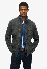 Mørkegrå jakke med knappede lommer, lynlåslukning og ståkrave, lagd over en blå denimskjorte. Båret med mørke jeans og et ur.