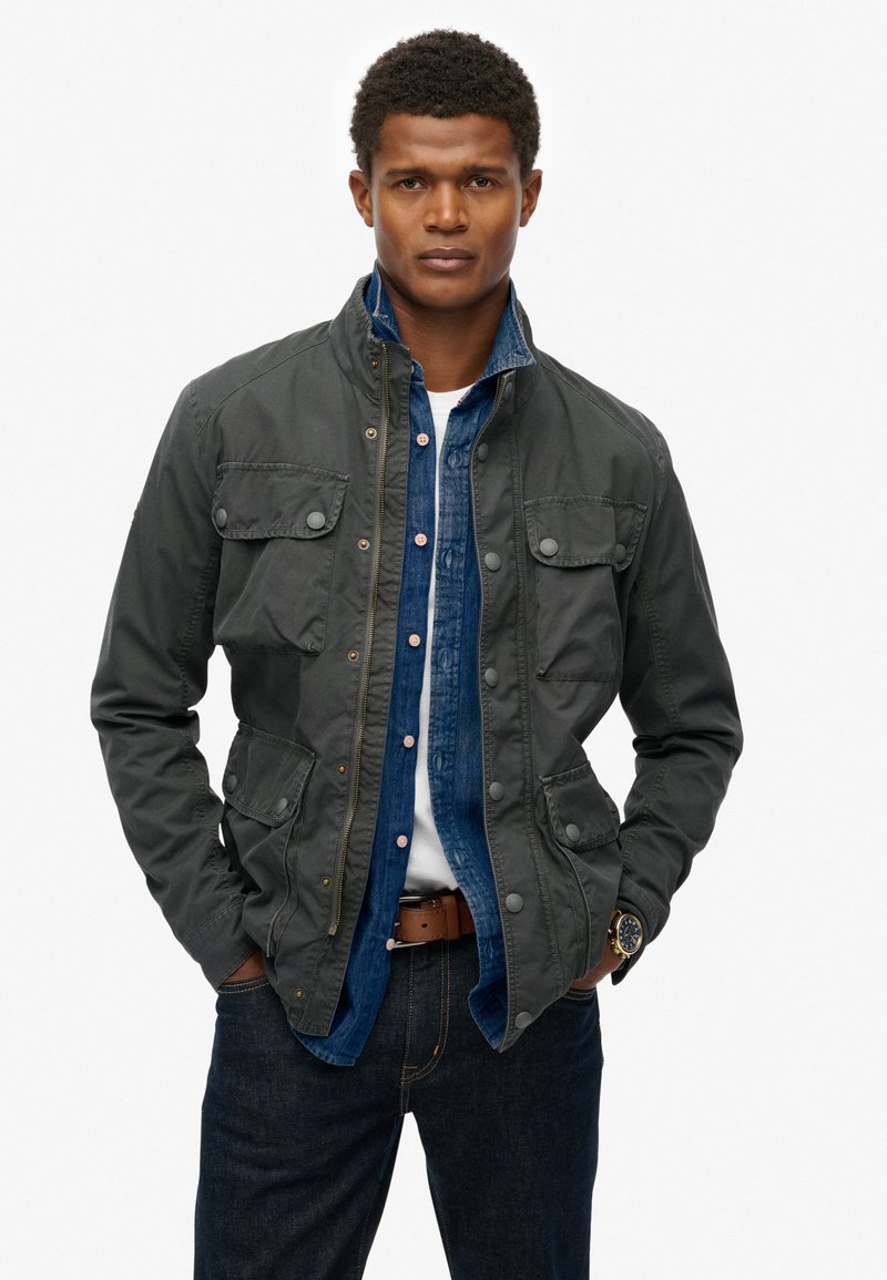 Mørkegrå jakke med knappede lommer, lynlåslukning og ståkrave, lagd over en blå denimskjorte. Båret med mørke jeans og et ur.