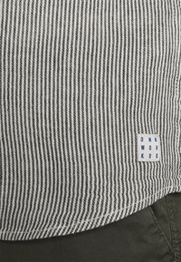 Gestreifter Stoff in Schwarz und Weiß mit gewebter Textur. Enthält ein blaues Etikett mit der Aufschrift "DNM WORKRS." Sichtbarer Rand eines grünen Stoffs darunter.