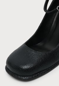 Gros plan d'une chaussure en cuir noir texturé avec un bout arrondi et une bride à la cheville sur fond blanc.