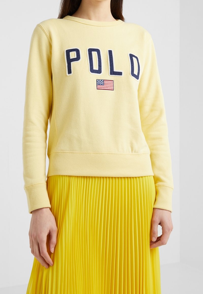 Persona che indossa una felpa giallo pallido con la scritta "POLO" e una bandiera americana ricamata, abbinata a una gonna plissettata gialla brillante.