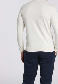 Homme portant un pull blanc à manches longues et un pantalon bleu foncé, debout, les mains partiellement dans les poches, tourné de dos sur un fond uni.