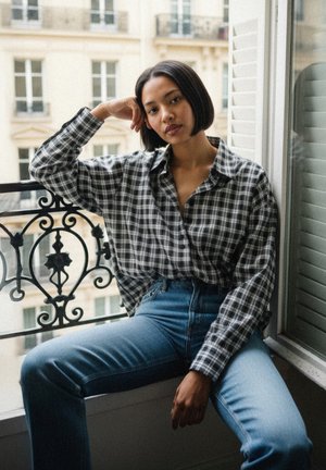 Black and white checkered button-up shirt paired with high-waisted blue jeans. Sitting by a window with decorative black iron railing.